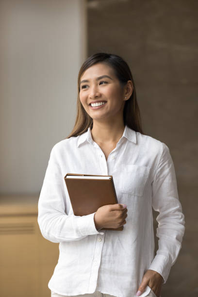 istock Cheerful young Asian student girl in white shirt holding notebook 1917884084