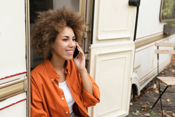 glückliche afrikanische junge reisende frau freiberuflerin telefoniert smartphone mit kunden verwandte freunde, entspannt reisen im van-anhänger wohnmobil, zuhause auf rädern - motor home mobile home isolated vehicle trailer stock-fotos und bilder