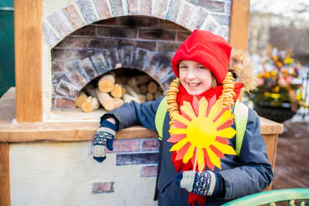 happy child at traditional russian festival maslenitsa, week of pancakes. the boy holds the sun in his hand, a symbol of the arrival of spring, stands by the stove at the fair.