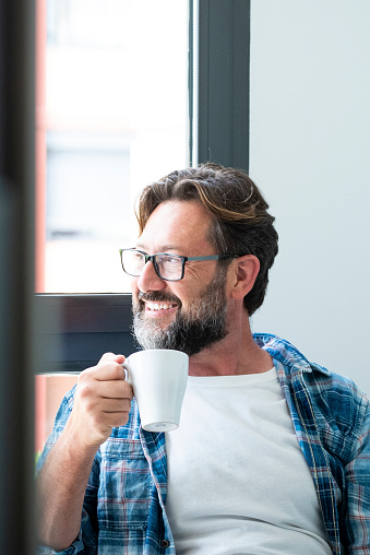 Portrait of happy man looking outside the window and smile drinking a coffee at home ot office - adult caucasian male with beard and glasses in job break activity alone - happy people with eyeglasses