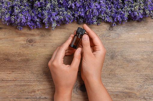 Woman with bottle of lavender essential oil and flowers on wooden background, top view