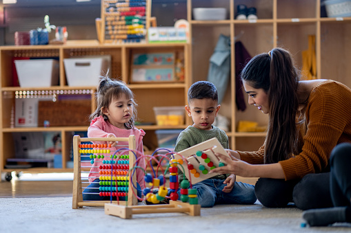 A kindergarten teacher sits on the floor with a small group of children as she encourages them in their play activities.  The children are each dressed casually and focused on tehir activities.