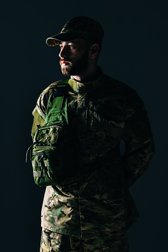 An army soldier with a bag over his shoulder, holds his hands behind his back in a dark room.