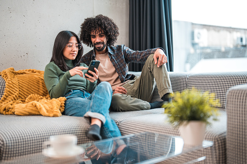 3/4 length image of an attractive Arab man sitting with both feet on a couch and his Asian female partner with legs crossed, enjoying the afternoon in  their chic apartment, both reading  savings plan offers on one smart phone and discussing. A coffee cup and flower pot on the glass table.