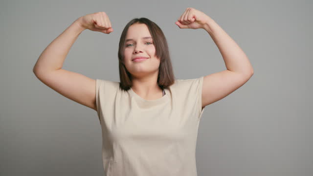 Young strong latin teenager girl feeling happy, satisfied and powerful, flexing fit and muscular biceps, looking strong on gray background