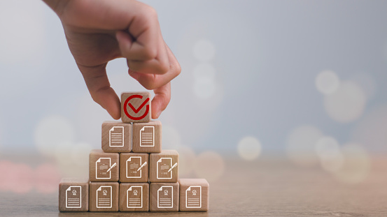 Hand-holding correct sign mark on wooden block cube and the top of document icon for ISO quality control certification and project approval concept.