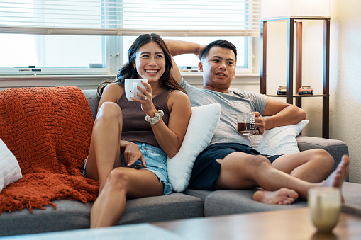 A beautiful Eurasian young woman of Pacific Islander descent relaxes on the sofa at home with her husband, a Pacific Islander young man who is enlisted in the USA Navy, as they watch TV and drink coffee.