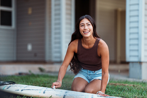 A beautiful Eurasian young woman of Hawaiian descent smiles as she waxes her surfboard in the front yard of her home before heading to the beach to surf.