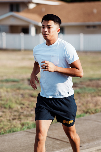 A fit Pacific Islander man who is enlisted in the Navy goes for a run along the street through his residential neighborhood in Hawaii on a sunny afternoon.