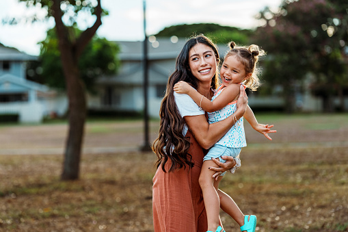 A cute Eurasian three year old girl laughs as her beautiful mom of Pacific Islander descent affectionately embraces her. The mom and daughter are having fun at the park together.