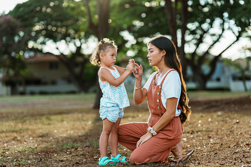 A loving mom of Pacific Islander descent kneels down to look at something her Eurasian three year old daughter found while playing outside at the park in their residential neighborhood.