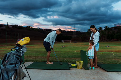 A cute and curious three year old girl of Pacific Islander descent affectionately holds hands with her mother and watches as her dad practices his golf swing at the driving range on a beautiful evening in Hawaii.