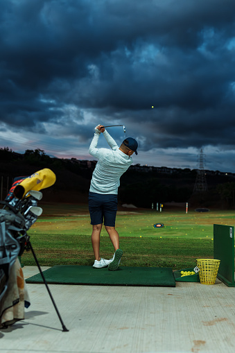 Rear view of a Pacific Islander male golf professional practicing his golf swing at the driving range on a cloudy evening in Hawaii.