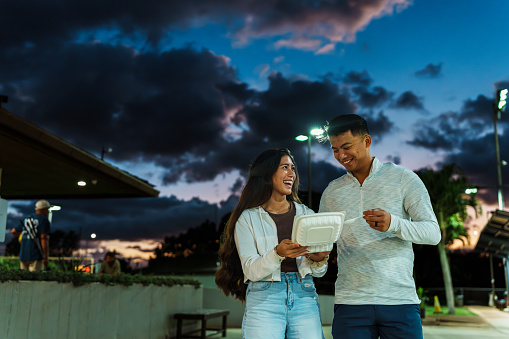 A loving young couple of Pacific Islander descent walk down the city street and share a take-out container of street food for dinner while on a date in Hawaii.