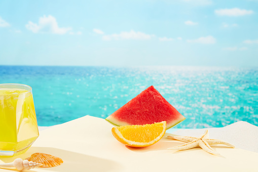 Juices, fresh fruits and shells are displayed on the white sand background, with the blue sea behind. Tropical atmosphere for advertising. Front view.