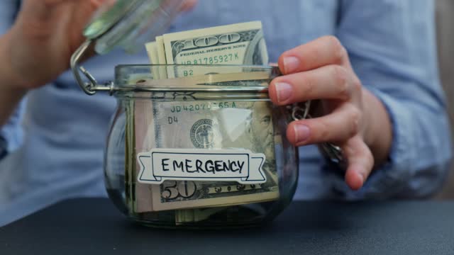 Unrecognizable woman turn Glass jar full of American currency dollars cash banknote with text EMERGENCY. Preparation saving money. Moderate consumption and economy