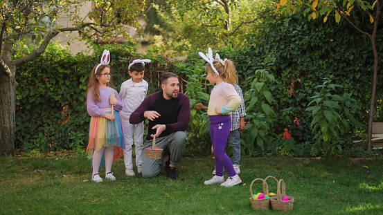 A young man is explaining the rules of egg and spoon race to a group of children outdoors.