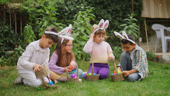 A group of children are sitting on grass and opening Easter eggs on Easter egg hunt game in a back yard garden.
