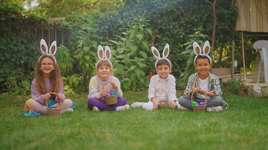 A portrait of children friends sitting on grass wearing bunny ears during Easter egg hunt in back yard garden.