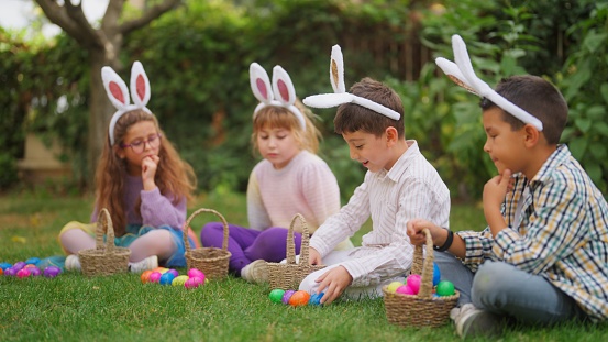 A group of children are sitting on grass and counting Easter eggs on Easter egg hunt game in a back yard garden.