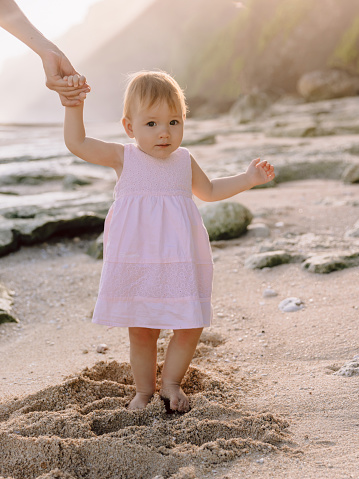Beautiful child girl staying in sand with sunset tones. Cute baby in pink dress on ocean coastline