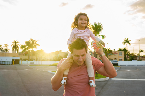 A loving and affectionate father of Caucasian descent carries his Eurasian toddler daughter on his shoulders during a walk outside at sunset.