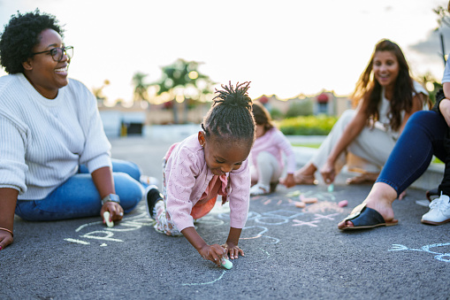 A toddler girl of African American descent draws with sidewalk chalk on the street in front of her home with friends and their moms.