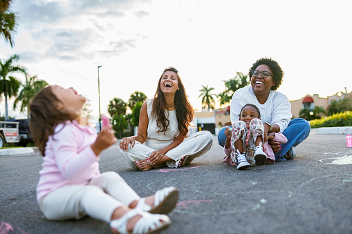 Two female adult best friends, one Asian and the other African American, laugh while coloring with sidewalk chalk with their toddler daughters.