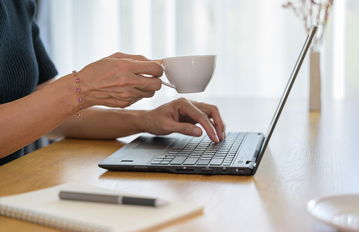 Close up of woman hand holding coffee cup while working at home with laptop in morning light