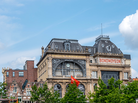 Jacob Van Artevelde statue in front of Socialist Bond Mouson historic building under blueish cloudscape. Green foliage and red flag.