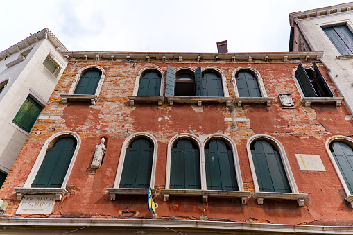 Old town of Venice with close-up of red weathered facade and windows with closed shutters on a blue cloudy summer day. Photo taken August 6th, 2023, Venice, Italy.
