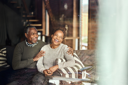 Happy African American mature couple enjoying on sofa in the living room. The view is through glass.