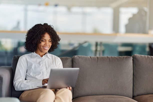 Happy black businesswoman using laptop on sofa in a hallway.