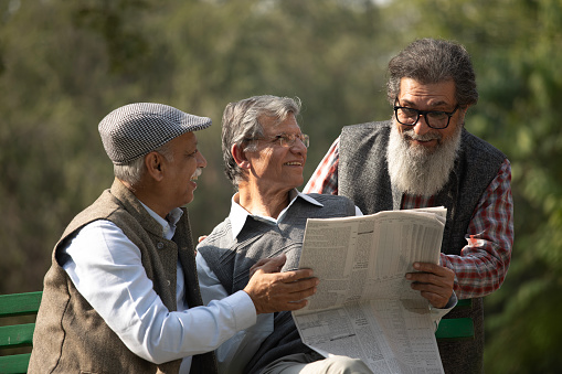 Elderly male friends smiling and reading newspaper on bench against trees at public park in morning