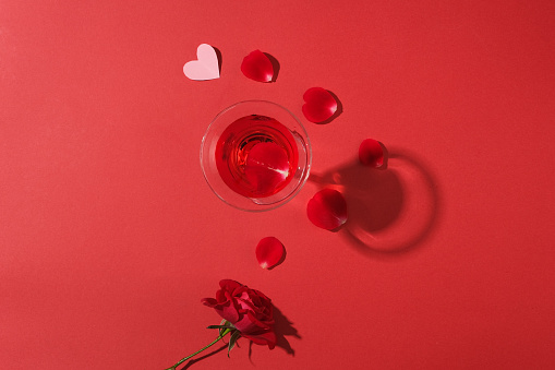 Red background featured a cocktail glass filled with red wine. A branch of rose and rose petals are decorated. Concept of Valentines Day for couples