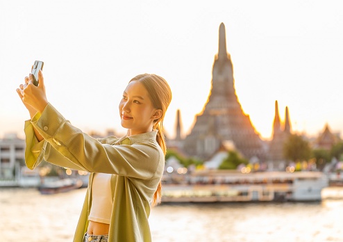 Embracing Sunset Serenity: Young Asian Woman Capturing Sunset Moments of Herself Against the Backdrop of Wat Arun in Bangkok Riverside, Thailand