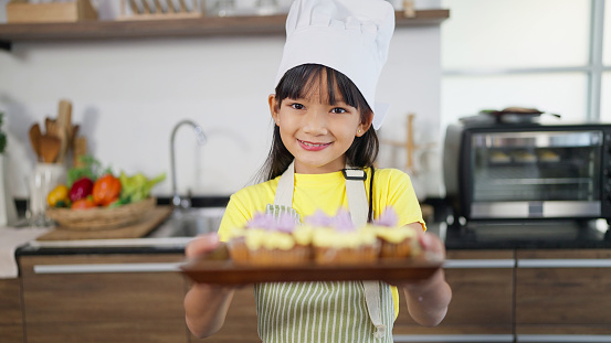 Portrait of smiling little asian girl in apron holding tray with freshly homemade cupcakes, muffins looking to camera. Concept for cooking learning for kid. Baking Concept