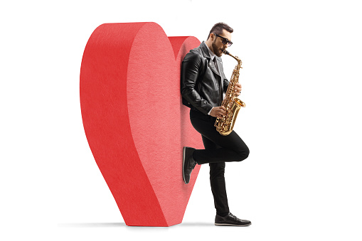 Full length profile shot of a man playing a saxophone and leaning on a red heart isolated on white background