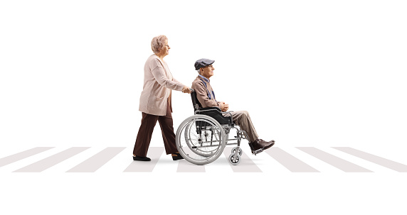 Full length profile shot of an elderly woman pushing an elderly man in a wheelchair at a pedestrian crossing isolated on white background