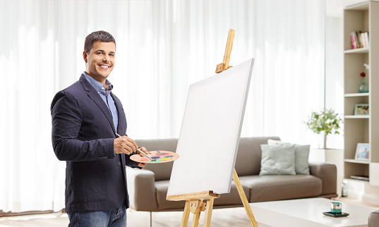 Young man painting on a canvas at home and smiling at camera