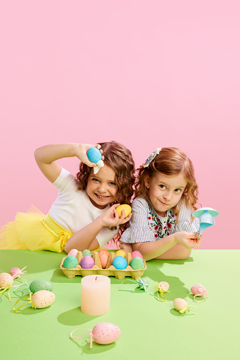 Beautiful little girls holding painted, decorated Easter eggs against pink background. Happiness. Concept of Easter holiday, celebration, traditions, childhood, happiness