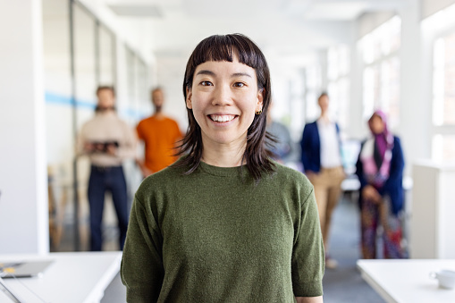 Close-up of smiling young Japanese businesswoman with team at back. Happy female executive standing in office with colleagues standing in the background.