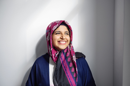 Portrait of a cheerful mid adult woman wearing headscarf leaning to wall looking away and smiling in office. Happe Islamic businesswoman standing in office.