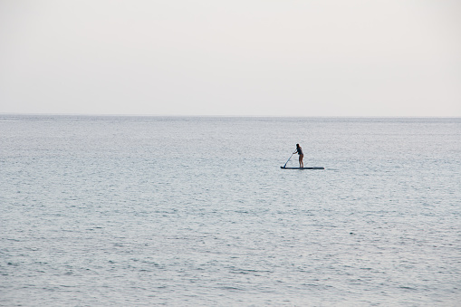 An unrecognizable woman in the distance, paddle surfing completely alone in a calm sea. Water sports concept