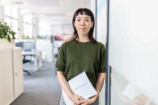 Portrait of Asian female professional looking at camera at office. Confident young woman standing in office holding a file.