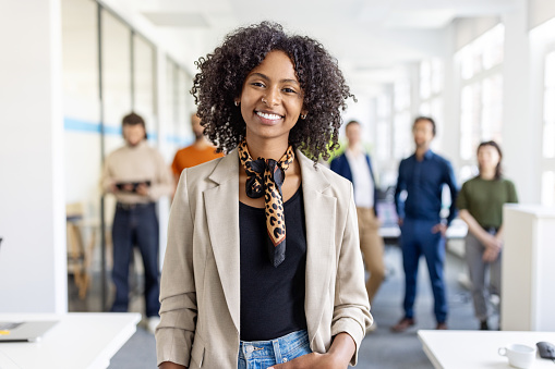 Portrait of young female entrepreneur with group of people at back. African businesswoman standing in office with group of business professionals in the background.
