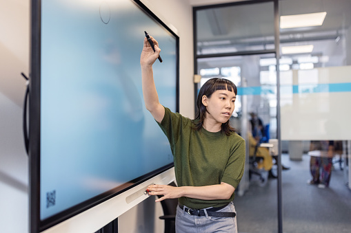 Young asian woman pointing at large television screen while giving presentation in office. Female entrepreneur giving presentation to team in office boardroom.