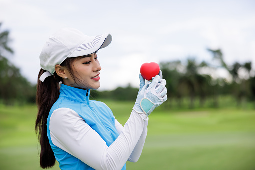 Portrait head shot of young asian female profestional golfer holding red heart at the course over white sky background,