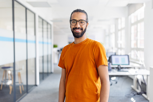 Portrait of happy young man standing in office. Smiling male entrepreneur with eyeglasses standing at startup looking at camera.