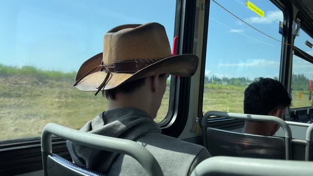 a man in a hat sits in a bus passing by a field cowboy headdress hooded sweatshirt a young man having fun going for a walk choosing clothes preference macho male style Canada Vancouver